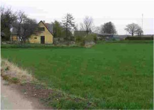 Foto: Oversigtsfoto. Brønd ligger til venstre for gul bygning mens hb1 ligger på opfyldt områder ved skel mellem græsplæne og mark. Hb2 ligger til højre for det gule hus ved skræntfoden af en lille skrænt mod grusvejen.